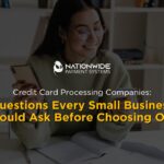 A small business owner smiling while looking closely at her credit card and using a smartphone to verify a transaction at a clean desk. An open notebook, a pen, and a ceramic cup of coffee sit in front of her. The white Nationwide Payment Systems logo is centered horizontally above bold yellow and white overlay text reading "Credit Card Processing Companies: Questions Every Small Business Should Ask Before Choosing One."