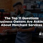 A professional workshop environment lifestyle photograph showing a focused business owner in a dark green t-shirt and protective denim work apron standing by a workbench filled with organized tools and blue parts bins. He is actively reviewing business metrics on his mobile smartphone with a cup of coffee nearby. Overlaid bold white text reads "The Top 11 Questions Business Owners Are Asking About Merchant Services," with the white Nationwide Payment Systems logo anchored cleanly in the bottom right corner.