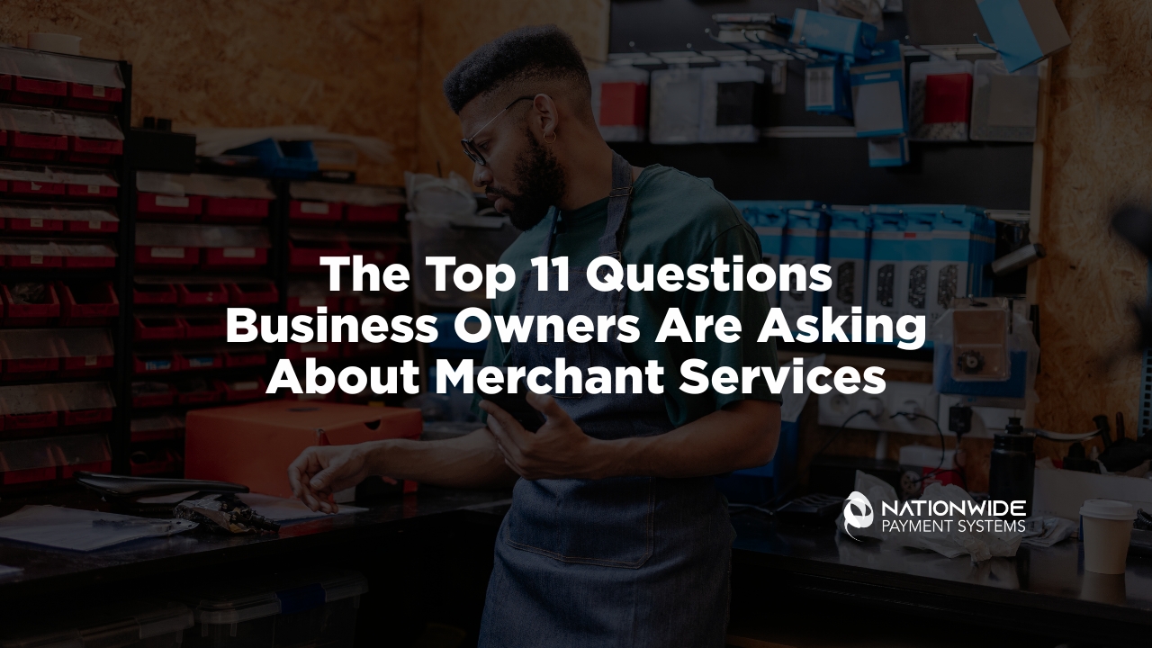 A professional workshop environment lifestyle photograph showing a focused business owner in a dark green t-shirt and protective denim work apron standing by a workbench filled with organized tools and blue parts bins. He is actively reviewing business metrics on his mobile smartphone with a cup of coffee nearby. Overlaid bold white text reads "The Top 11 Questions Business Owners Are Asking About Merchant Services," with the white Nationwide Payment Systems logo anchored cleanly in the bottom right corner.