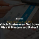A hand holding a pen pointing to numbers on a printed financial chart alongside a desk calculator and laptop. Centered on the image is the Nationwide Payment Systems logo above yellow subtext reading "Special Interchange Rates in 2026:" and bold white text reading "Which Businesses Get Lower Visa & Mastercard Rates?"
