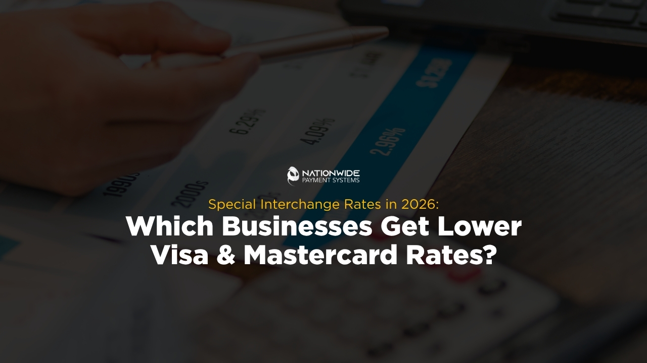 A hand holding a pen pointing to numbers on a printed financial chart alongside a desk calculator and laptop. Centered on the image is the Nationwide Payment Systems logo above yellow subtext reading "Special Interchange Rates in 2026:" and bold white text reading "Which Businesses Get Lower Visa & Mastercard Rates?"